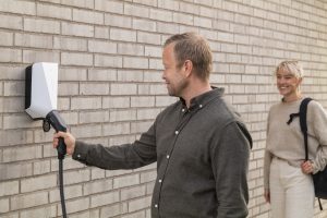 A man using a black Easee charger mounted on a brick wall to charge a polestar. A woman watches in the background.