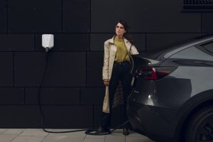 White Easee charger mounted on dark wall. Woman charging a Tesla Model Y.